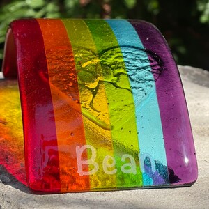 Puede incluir: Una pieza de arte de vidrio fusionado de color arco&iacute;ris con el nombre "Bean" grabado en el vidrio. La pieza tiene forma curva, con los colores del arco&iacute;ris dispuestos en franjas verticales. Un dise&ntilde;o de coraz&oacute;n y hoja est&aacute; grabado.