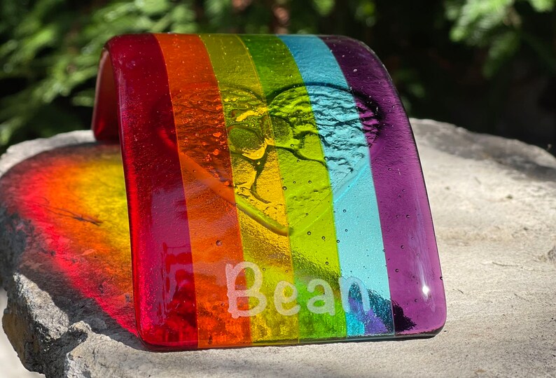 Puede incluir: Una pieza de arte de vidrio fusionado de color arco&iacute;ris con el nombre "Bean" grabado en el vidrio. La pieza tiene forma curva, con los colores del arco&iacute;ris dispuestos en franjas verticales. Un dise&ntilde;o de coraz&oacute;n y hoja est&aacute; grabado.