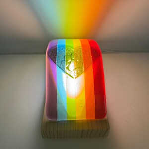 Puede incluir: Una luz de noche de cristal con los colores del arco&iacute;ris y un dise&ntilde;o en forma de coraz&oacute;n. La luz est&aacute; montada sobre una base de madera y proyecta un arco&iacute;ris en la pared. El cristal presenta rayas verticales con los colores del arco&iacute;ris.