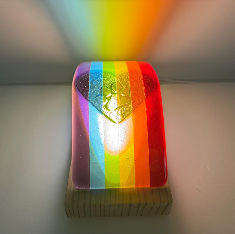Puede incluir: Una luz de noche de cristal con los colores del arco&iacute;ris y un dise&ntilde;o en forma de coraz&oacute;n. La luz est&aacute; montada sobre una base de madera y proyecta un arco&iacute;ris en la pared. El cristal presenta rayas verticales con los colores del arco&iacute;ris.