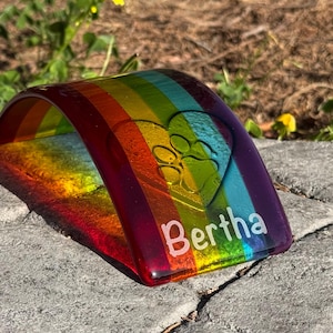 Puede incluir: Arco conmemorativo para mascotas de vidrio colorido con dise&ntilde;o de arco&iacute;ris. El arco presenta una huella de pata dentro de un coraz&oacute;n y el nombre "Bertha" en letras blancas. Colocado sobre una superficie de piedra.