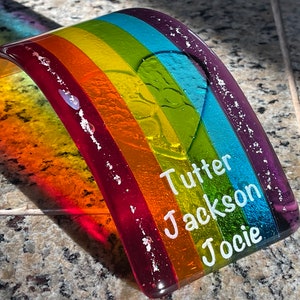 Puede incluir: Un arco&iacute;ris de cristal colorido con los nombres "Tutter", "Jackson" y "Jocie" en letras blancas. El arco&iacute;ris presenta bandas rojas, naranjas, amarillas, verdes, azules y moradas. El objeto de cristal curvado se muestra sobre una superficie moteada.