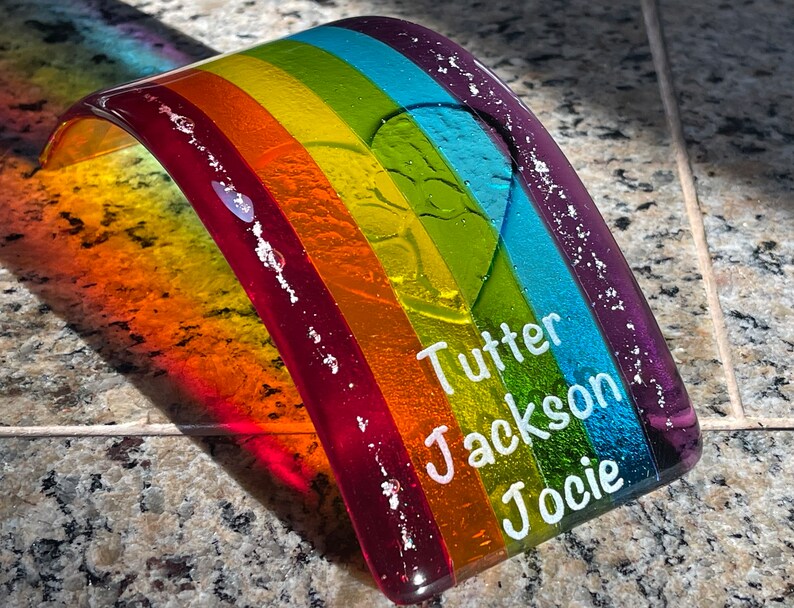 Puede incluir: Un arco&iacute;ris de cristal colorido con los nombres "Tutter", "Jackson" y "Jocie" en letras blancas. El arco&iacute;ris presenta bandas rojas, naranjas, amarillas, verdes, azules y moradas. El objeto de cristal curvado se muestra sobre una superficie moteada.