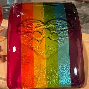Puede incluir: Una pieza de arte de vidrio rectangular con un dise&ntilde;o de rayas de arco&iacute;ris. El vidrio presenta una forma de coraz&oacute;n con huellas de patas. Los colores incluyen rojo, naranja, amarillo, verde, azul y morado. Una granada es visible en el fondo.