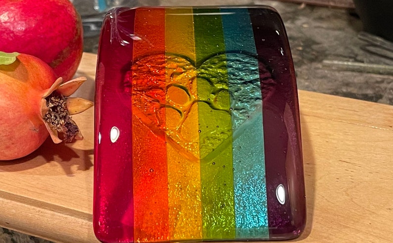 Puede incluir: Una pieza de arte de vidrio rectangular con un dise&ntilde;o de rayas de arco&iacute;ris. El vidrio presenta una forma de coraz&oacute;n con huellas de patas. Los colores incluyen rojo, naranja, amarillo, verde, azul y morado. Una granada es visible en el fondo.
