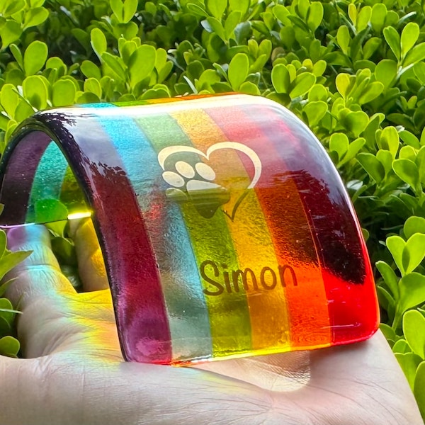 Pode incluir: Uma peça de vidro curva e retangular com listras verticais nas cores do arco-íris, incluindo vermelho, laranja, amarelo, verde, azul e roxo. O vidro é gravado com um coração contendo uma pegada e o nome Simon em fonte sem serifa.