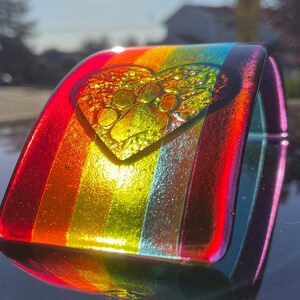Puede incluir: Una pulsera de vidrio con los colores del arco&iacute;ris, con un dije en forma de coraz&oacute;n con un dise&ntilde;o de huella de pata. La pulsera tiene una forma curva y un acabado brillante, que refleja la luz. Los colores van del rojo al amarillo y al verde.
