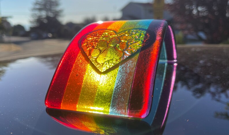 Puede incluir: Una pulsera de vidrio con los colores del arco&iacute;ris, con un dije en forma de coraz&oacute;n con un dise&ntilde;o de huella de pata. La pulsera tiene una forma curva y un acabado brillante, que refleja la luz. Los colores van del rojo al amarillo y al verde.