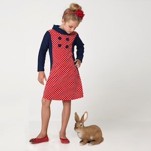 May include: A young girl wearing a red and blue polka dot dress with a navy blue collar and three buttons. She is wearing a red flower headband. A brown rabbit figurine is in the foreground.