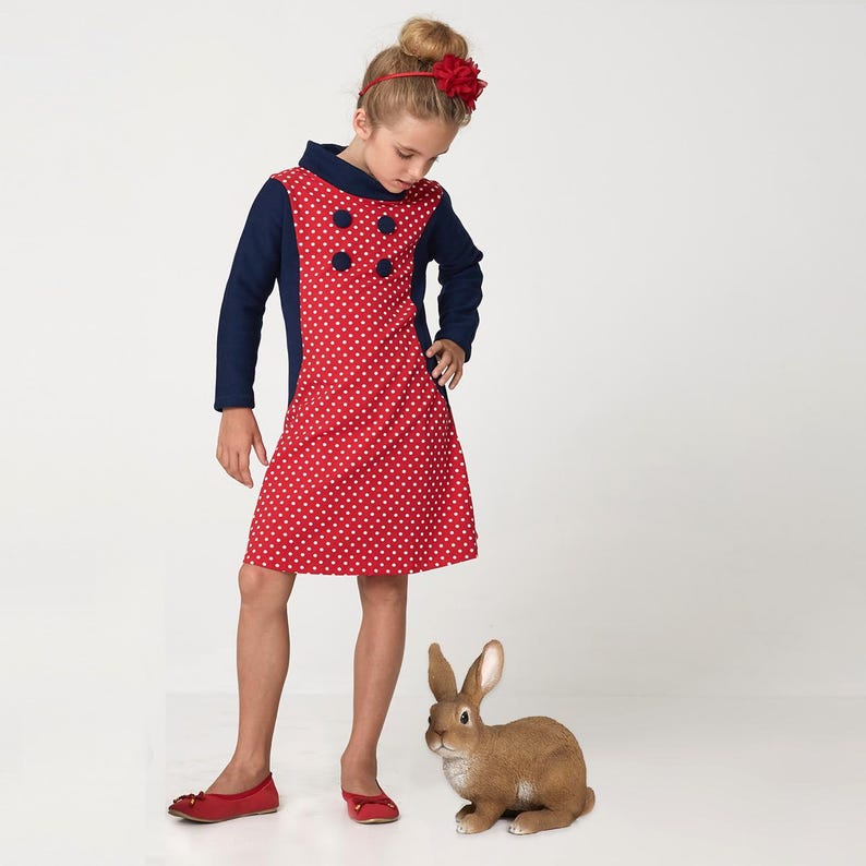 May include: A young girl wearing a red and blue polka dot dress with a navy blue collar and three buttons. She is wearing a red flower headband. A brown rabbit figurine is in the foreground.