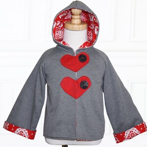 Op de afbeelding: Een grijze hoodie met rode hartvormige patches en zwarte knopen. De mouwen en de capuchon zijn gevoerd met een rood-witte stof met een patroon.