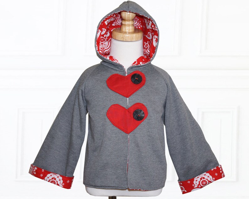 Op de afbeelding: Een grijze hoodie met rode hartvormige patches en zwarte knopen. De mouwen en de capuchon zijn gevoerd met een rood-witte stof met een patroon.