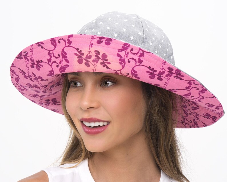 Puede incluir: Un sombrero de sol de ala ancha con un estampado floral rosa en la parte inferior del ala y una tela gris con estrellas blancas en la parte superior. El sombrero lo lleva una persona con el pelo largo y casta&ntilde;o.