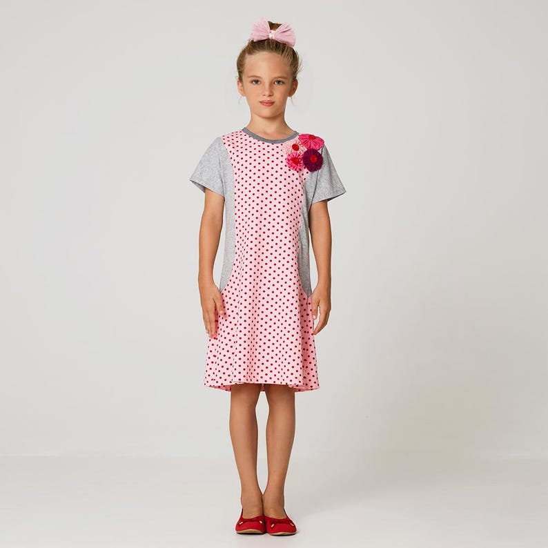 May include: A pink and grey polka dot dress with a flower embellishment on the shoulder. The dress has short sleeves and a slightly flared skirt.