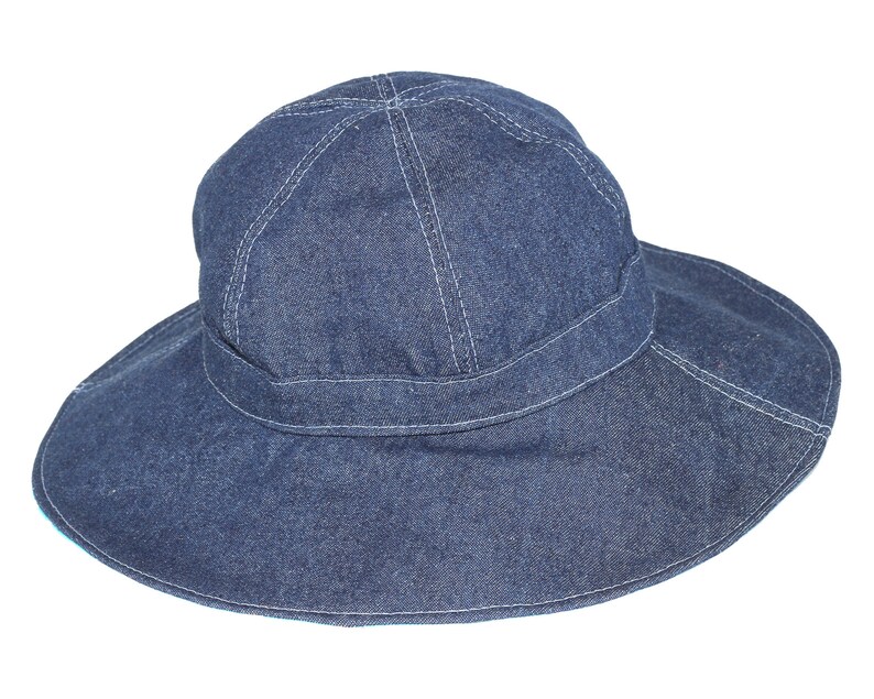 Puede incluir: Un sombrero de sol de denim de ala ancha de color azul oscuro. El sombrero tiene una banda cosida alrededor de la corona y un ala ancha.