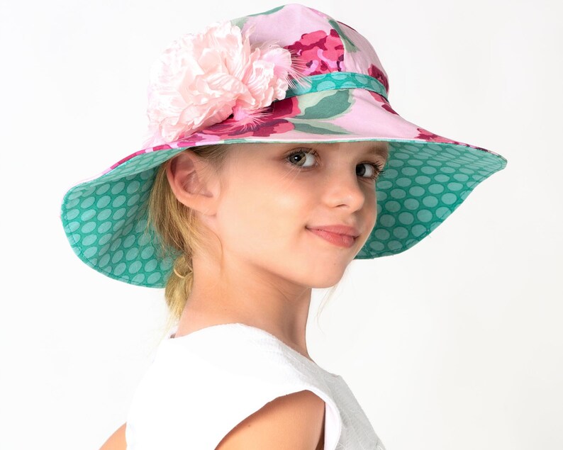 Puede incluir: Un sombrero de sol floral rosa y verde con una gran flor rosa en el lateral. El sombrero tiene un forro de lunares.