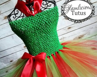 May include: A green and red tutu with a green crocheted top and a red satin bow. The tutu is made of tulle and has a layered design. The top is sleeveless and has a halter neckline. The bow is tied at the waist. The tutu is perfect for a holiday party or special occasion.