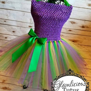 May include: Purple and green tulle tutu with a purple sequined bodice and a green satin bow. The tutu has layers of tulle in purple, green, and yellow. The bodice is made of a stretchy fabric with sequins. The bow is tied around the waist. The tutu is perfect for a Mardi Gras celebration.