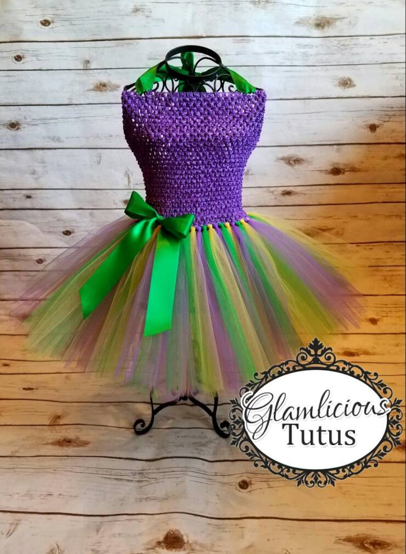 May include: A purple and green tulle tutu dress with a green satin bow. The dress is on a black stand against a wood background. The text "Glamlicious Tutus" is visible in the bottom right corner of the image.
