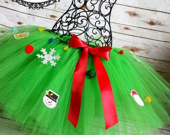 May include: A green tulle tutu decorated with Christmas-themed embellishments, including a snowman, snowflake, and ornaments. A red satin ribbon is tied around the waist.