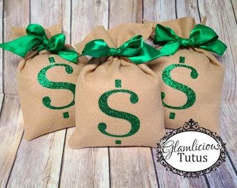 May include: Three tan fabric money bags with green glitter dollar signs and green satin bows. The bags are on a wooden surface. The bags are part of a set.