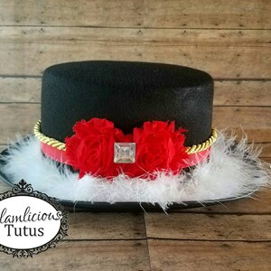 Pu&ograve; includere: Un cappello a cilindro nero con un bordo dorato e fiori di stoffa rossi. Il cappello &egrave; decorato con piume bianche.