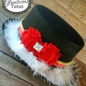 Pu&ograve; includere: Cappello a cilindro nero con una fascia dorata, decorazioni floreali rosse e bianche e un bordo di piume bianche. Il cappello ha un strass trasparente al centro dei fiori.