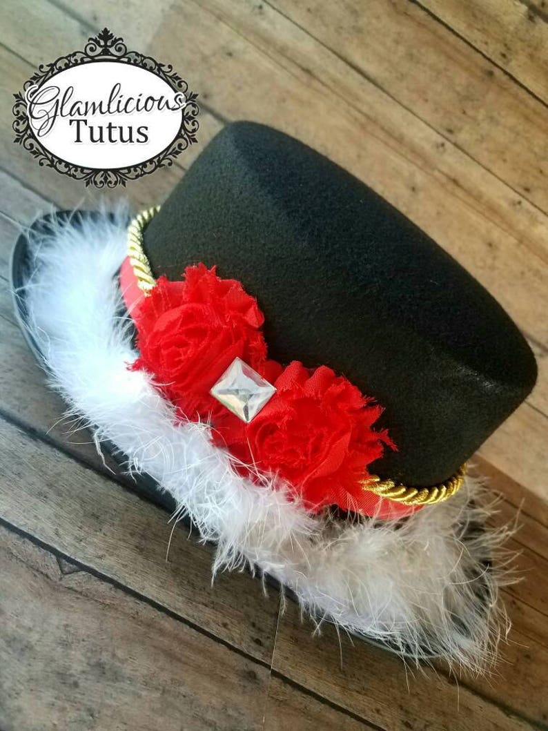 Pu&ograve; includere: Cappello a cilindro nero con una fascia dorata, decorazioni floreali rosse e bianche e un bordo di piume bianche. Il cappello ha un strass trasparente al centro dei fiori.