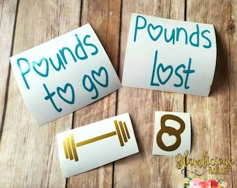 May include: Four turquoise and gold vinyl decals with the words "Pounds to go", "Pounds Lost", a dumbbell, and a kettlebell. The decals are on a wood background.