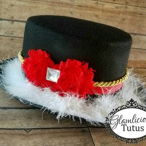 Pu&ograve; includere: Un cappello a cilindro nero con un bordo dorato e fiori di tessuto rosso e bianco. Il cappello &egrave; decorato con un strass trasparente e ha un bordo di piume bianche.