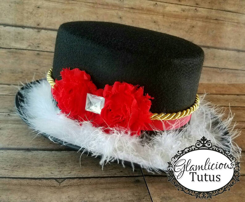 Pu&ograve; includere: Un cappello a cilindro nero con un bordo dorato e fiori di tessuto rosso e bianco. Il cappello &egrave; decorato con un strass trasparente e ha un bordo di piume bianche.