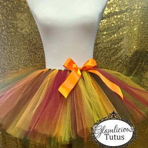 May include: A multi-colored tulle tutu with an orange satin bow. The tutu is made of layers of tulle in shades of red, orange, yellow, and green. The tutu is on a white mannequin. The text "Glamlicious Tutus" is visible in the lower right corner of the image.
