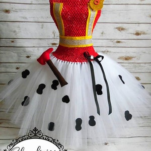 May include: A white tulle tutu with black polka dots and a black ribbon. The tutu is paired with a red and yellow bodice with a silver trim and a gold badge with the number "8" on it. A red and brown toy axe is included.