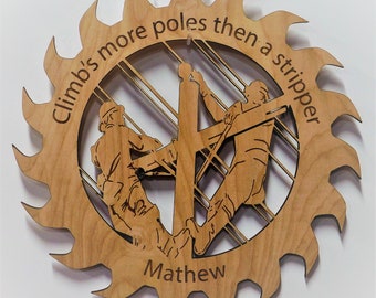 May include: A wooden saw blade shaped plaque with a silhouette of two people climbing a utility pole. The plaque reads "Climb's more poles then a stripper" and is personalized with the name "Mathew".