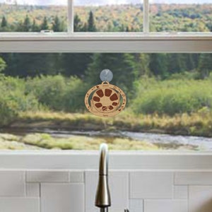 May include: A wooden ornament with a circular design featuring a sun with rays and a mountain range. The ornament is brown with a red sun and is labeled "2023" and "Home".