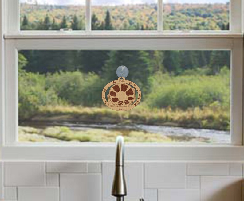 May include: A wooden ornament with a circular design featuring a sun with rays and a mountain range. The ornament is brown with a red sun and is labeled "2023" and "Home".