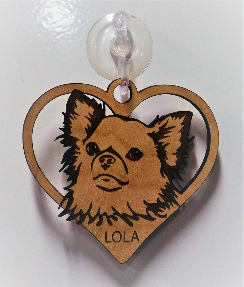 May include: A wooden heart-shaped ornament with a cutout of a chihuahua dog's head. The dog's fur is brown and black, and the ornament is inscribed with the name "LOLA".