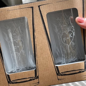May include: Two clear glass shot glasses with a tree design and the initials "F+K" etched into a heart shape. The glasses are in a brown cardboard box.