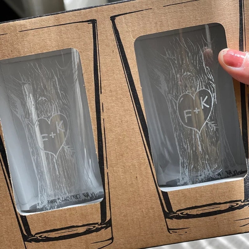 May include: Two clear glass shot glasses with a tree design and the initials "F+K" etched into a heart shape. The glasses are in a brown cardboard box.