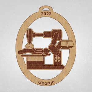 May include: A brown wooden ornament shaped like a circle with a brown silhouette of a person lying on a table with a medical device above them. The ornament has the year 2022 and the name "George" inscribed on it.