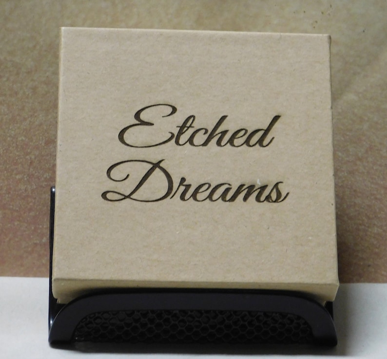 May include: A brown cardboard box with the words "Etched Dreams" written in black cursive lettering. The box is sitting on a black metal stand.