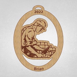 May include: Wooden ornament featuring a construction worker laying bricks, with the year 2022 at the top. The name "Brian" is at the bottom. The ornament is oval-shaped with a hole for hanging.