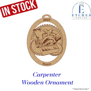 May include: A brown wooden ornament shaped like a circle with a carpenter using a saw to cut wood. The ornament has the year 2023 and the word "Name" written on it. The ornament is a Christmas decoration.