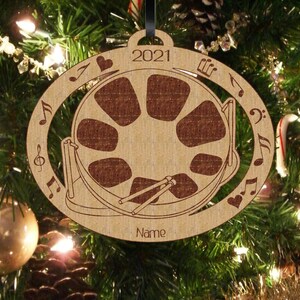 May include: A wooden Christmas ornament shaped like a steel drum with musical notes and a heart around the edge. The ornament is brown and has the year 2021 and the word "Name" on it.
