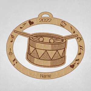 May include: A brown wooden Christmas ornament shaped like a drum with musical notes and a heart around the edge. The drum has a brown and white striped pattern and the text "2022" and "Name" are printed on the ornament.