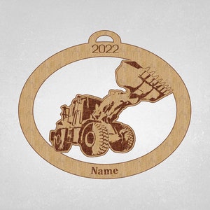 May include: A brown wooden Christmas ornament shaped like a circle with a cutout of a front loader excavator. The ornament has the year 2022 and the word "Name" printed on it.