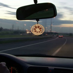 May include: A wooden car charm with a circular design featuring a brown and white mandala pattern. The charm is hanging from the rearview mirror of a car.