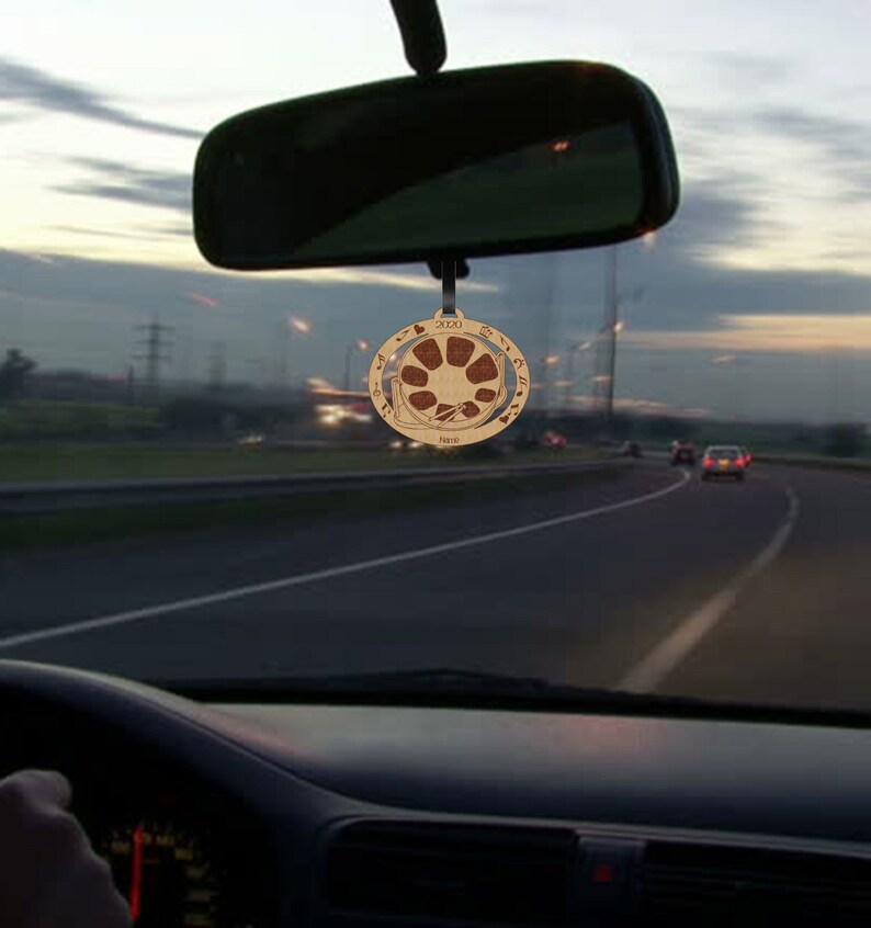 May include: A wooden car charm with a circular design featuring a brown and white mandala pattern. The charm is hanging from the rearview mirror of a car.