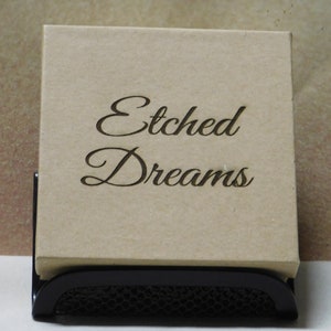 May include: A brown cardboard box with the words "Etched Dreams" written in black cursive lettering. The box is sitting on a black metal stand.