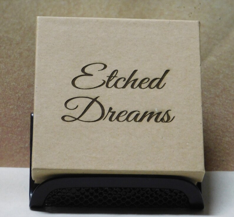 May include: A brown cardboard box with the words "Etched Dreams" written in black cursive lettering. The box is sitting on a black metal stand.